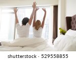 Back view of young happy couple in white t-shirts waking up in the morning, sitting on bed, stretching in cozy bedroom, looking through window at big city scenery. Funny married couple after wakeup