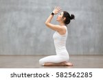 Beautiful young woman wearing sport watch for heart rate and calorie burn control working out against grey wall, doing yoga or pilates exercise. Girl sitting in seiza, vajrasana pose. Full length 