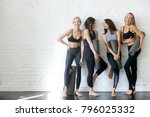 Group of young sporty girls with yoga mats standing at white wall. Female companions in gym resting after fitness, indoor full length, loft studio, copy space