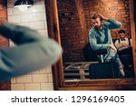 Small photo of Barbershop concept. Reflection of red bearded harsh stylish man in the mirror. He has a fashionable hairstyle, modern haircut, wearing jeans casual outfit, waiting for a hairstylist