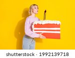 Photo of cheerful nice amazed young lady hold hands big slice cake blow candle isolated on yellow color background