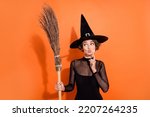 Small photo of Portrait of charming witch person finger touch chin pouted lips look empty space hold broom isolated on orange color background
