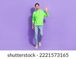 Full length photo of funny cheerful man wear green hoodie walking waving arm isolated purple color background
