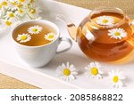 Healthy herbal tea in cup, chamomile flowers and teapot on white wooden tray.