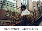 Small photo of Young smiling african woman using smartphone going downstairs in the city