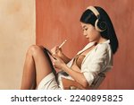 Profile view of asian woman in headphones drawing on tablet with stylus pen, sitting on chair, graphic designer working as freelancer, creating digital art
