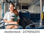 Small photo of Attractive young adult woman listening music while traveling by city shuttle bus, holding travel mug in hands.