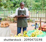 Happy African male trader or vendor holding a smart phone while standing at his fruit stall in a market place