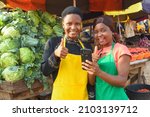 Small photo of Two happy African business women or female traders wearing colorful aprons while standing at a vegetable stall in a market place with a smart phone with them