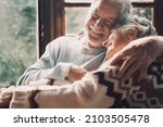 Small photo of Old senior couple in love hug and embrace with romance together at home with outside view in windows background - happy mature retired people lifestyle enjoying caring each other and smiling