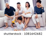 Small photo of Happy Asian extended family sitting on sofa together and watching little child playing toy on the floor with happiness