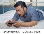 Small photo of Young Asian plus size man playing a video game on mobile phone and got negative reactions on bed at home. Facial expressions could be furious, fierce, upset, unhappy, irritated, frustrated.