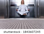 Horizontal low angle shot of a businesswoman meditating sitting cross-legged levitating in an open elevator in office with copy space.