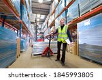 A wide shot of a female warehouse manager holding a clipboard and pulling a pallet truck.