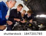 Small photo of A side view of a young auto mechanics student repairing a car while being observed by his trainer and other students in an automotive training school