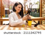 Brunette Caucasian business lady with small wireless black headphones in her ears looks at the phone with a smile, reading messages. Woman is relaxing in a cafe on the terrace enjoying music