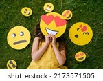 The person is lying on the grass covering his face with a loving emoticon with big hearts instead of eyes. Smile faces with different moods next to the child