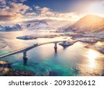 Aerial view of bridge, sea and snowy mountains in Lofoten Islands, Norway. Fredvang bridges at sunset in winter. Landscape with blue water, rocks in snow, road and sky with clouds. Top view from drone