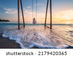 Happy young woman on wooden swing in water, beautiful blue sea with waves, sandy beach, golden sky at sunset. Summer holiday in Oludeniz, Turkey. Girl ride on a swing on sea coast, clear water. Travel