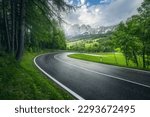 Road in green forest in rainy summer day. Dolomites, Italy. Beautiful mountain roadway, tress, grass, high rocks, blue sky with clouds. Landscape with empty highway through the wood in spring. Travel	