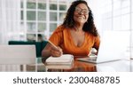 Small photo of Black woman happily planning her retirement from the comfort of her own home. Mature woman sitting at a table with her laptop, writing her goals and dreams for the future in a journal.
