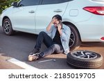Small photo of Handsome Asian man checking the punctured tyre on his car loosening the nuts with a wheel spanner before jacking up the vehicle in street. Men worry and call to service car. Change tire.
