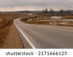 Low angle photo of tarmac curvy road at vintage brown meadows at early spring. Melancholic winding drive at countryside from left. Twisty gray asphalt path near Ergli, Latvia during rainy day. 