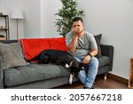 Small photo of Young latin man and dog sitting on the sofa at home smelling something stinky and disgusting, intolerable smell, holding breath with fingers on nose. bad smell