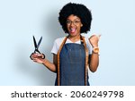 Small photo of Young african american woman dressmaker designer wearing atelier apron holding scissors pointing thumb up to the side smiling happy with open mouth