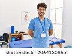 Small photo of Young hispanic man wearing physio therapist uniform standing at clinic