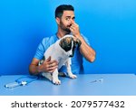 Small photo of Handsome hispanic veterinary man with beard checking dog health smelling something stinky and disgusting, intolerable smell, holding breath with fingers on nose. bad smell