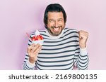 Middle age caucasian man eating strawberry ice cream screaming proud, celebrating victory and success very excited with raised arm 