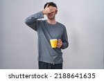 Small photo of Young hispanic man wearing pajama drinking a cup of coffee covering eyes with hand, looking serious and sad. sightless, hiding and rejection concept