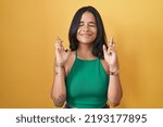 Small photo of Brunette woman standing over yellow background gesturing finger crossed smiling with hope and eyes closed. luck and superstitious concept.