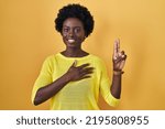 Small photo of African young woman standing over yellow studio smiling swearing with hand on chest and fingers up, making a loyalty promise oath