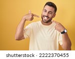 Small photo of Handsome hispanic man standing over yellow background smiling cheerful showing and pointing with fingers teeth and mouth. dental health concept.