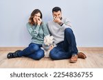 Small photo of Young hispanic couple sitting on the floor with dog smelling something stinky and disgusting, intolerable smell, holding breath with fingers on nose. bad smell