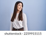 Small photo of Young brunette woman standing over blue background looking away to side with smile on face, natural expression. laughing confident.