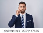 Small photo of Handsome hispanic man wearing suit and tie mouth and lips shut as zip with fingers. secret and silent, taboo talking