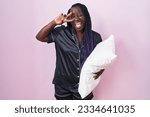 Young african woman wearing pijama hugging pillow doing peace symbol with fingers over face, smiling cheerful showing victory 