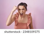 Small photo of Young teenager girl standing over pink background angry and mad raising fist frustrated and furious while shouting with anger. rage and aggressive concept.