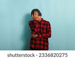 Small photo of A frustrated young Asian man with a beanie hat and a red plaid flannel shirt keeps his hand on his forehead, filled with regret for his wrongdoings, and feels stressed, isolated on blue background