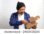 Small photo of An excited Asian man, dressed in a beanie hat and casual shirt, eagerly unpacks the cardboard parcel package, delightfully unboxing his awaited online store delivery from an online shopping spree