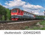 A red electric locomotive with a freight train moves along the railroad tracks. Close-up. Freight train on the way next to the green forest against the blue sky. Russia, Ural
