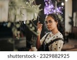 an Asian woman in glasses and wearing overalls standing near a hanging plant in front of a coffee shop terrace at night
