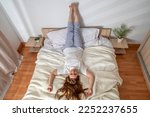 Woman doing exercises in bed at home in the morning. Yoga pose Viparita Karani or legs up the wall body stretch. Lady with long hair in pajamas. Girl stretches her muscles on a clean white bedding
