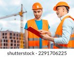 Small photo of Older construction supervisor with younger worker helper at construction site. Managers wearing protective workwear, hard hat, construction cranes on skyline. Construction workforce, working labor man