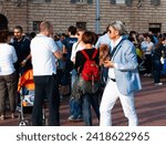 Small photo of Gubbio Umbria, Italy-May 15 2011; Stylish male dressed in light blue and carrying a dog walks past People gathered in European town square as sun lowers and shadows lengthen in late afternoon.