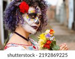 outdoor portrait of young latin caucasian woman, with face painted as La Calavera catrina, standing on blurred cemetery in background and copy space.