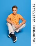 Small photo of Young man in orange undershirt and white sweatpants sits on studio floor over blue background
